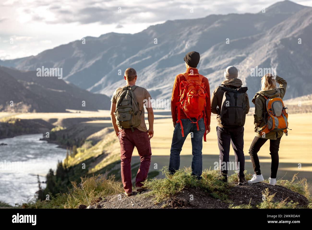 Eine Gruppe junger Wanderer mit Rucksäcken steht am Aussichtspunkt in den Bergen und genießt einen schönen Blick auf den Sonnenuntergang und den großen Fluss Stockfoto