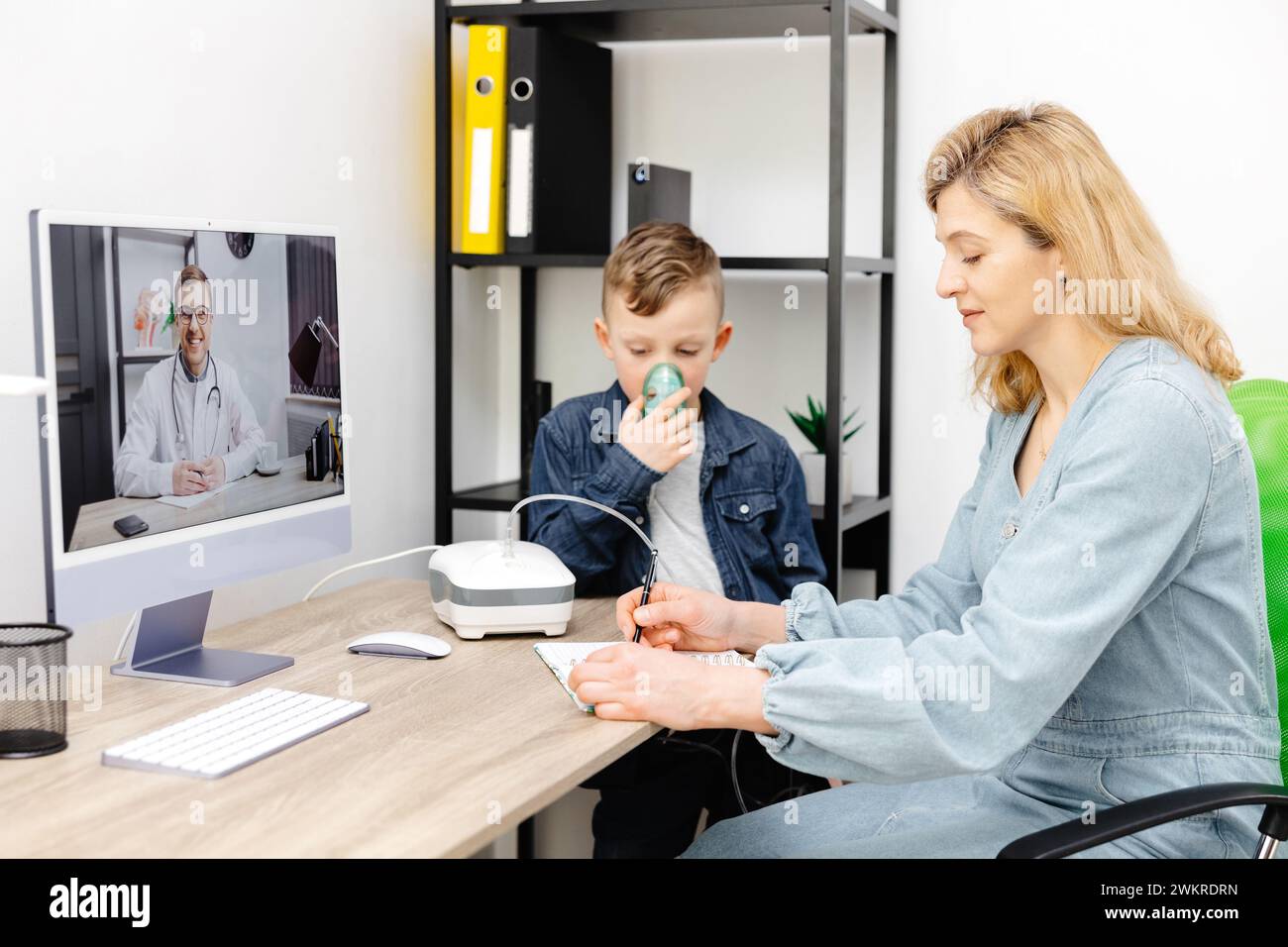 Kaukasischer kleiner Junge, der zu Hause mit einem Vernebler inhaliert. Eine junge Mutter, die ihrem Sohn beim Einatmen mit einem Vernebler hilft, bei einem Online-Meeting mit einem D. Stockfoto