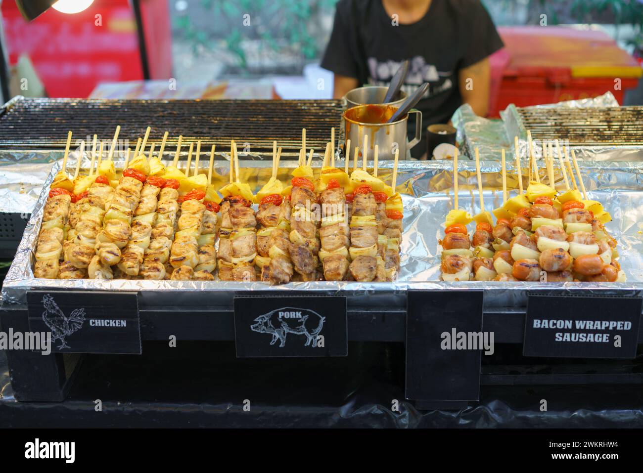 Eine appetitliche Auswahl an gegrillten Spießen mit Hühnchen, Schweinefleisch und mit Speck gewickelten Würstchen, begleitet von Gemüse, wird in einem Street Food stan präsentiert Stockfoto