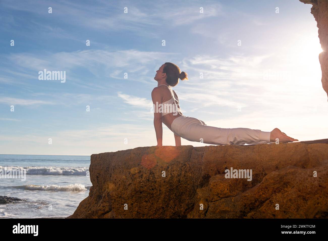 Eine Frau, die bei Sonnenaufgang auf einem Felsen am Meer eine nach oben gerichtete Hundeschuga-Pose macht. Stockfoto