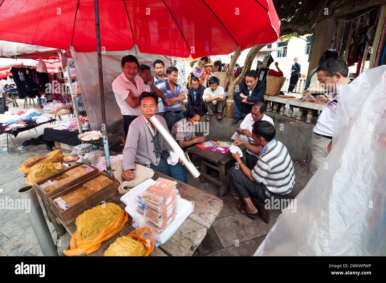 Ein Mann, der eine Triditionspfeife raucht, umgeben von einer Gruppe Männer, die Karten spielen, an einem Verkaufsstand im ländlichen Yunnan China Stockfoto