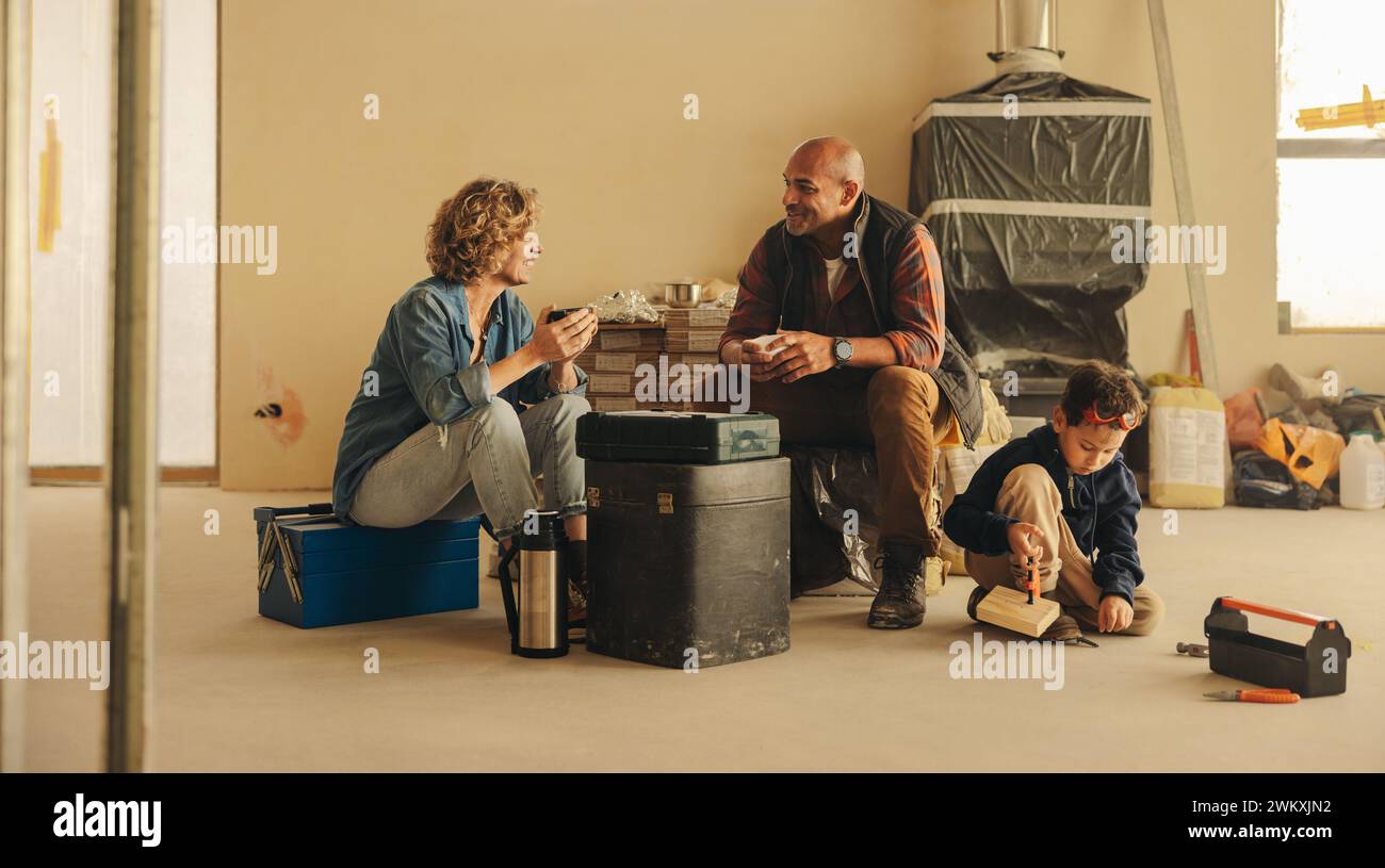 Das Paar und ihr kleiner Sohn machen eine Pause von der Renovierung ihres Hauses. Zusammen sitzen sie drinnen und genießen Kaffee, während sie sich für ihr DIY-Projekt aufladen Stockfoto