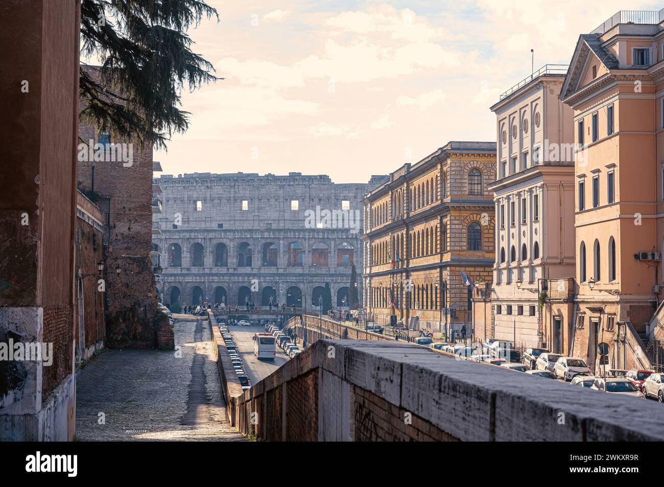 Malerische Straße in Rom, Italien. Das berühmte Kolosseum ragt in der Ferne und taucht in der Morgensonne auf. Sonnenstrahlen beleuchten das nahe gelegene b Stockfoto