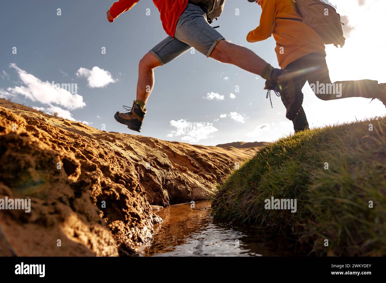 Zwei aktive junge Touristen Wanderer mit Rucksäcken springen über einen Bach. Gras- und Sandbachufer Stockfoto