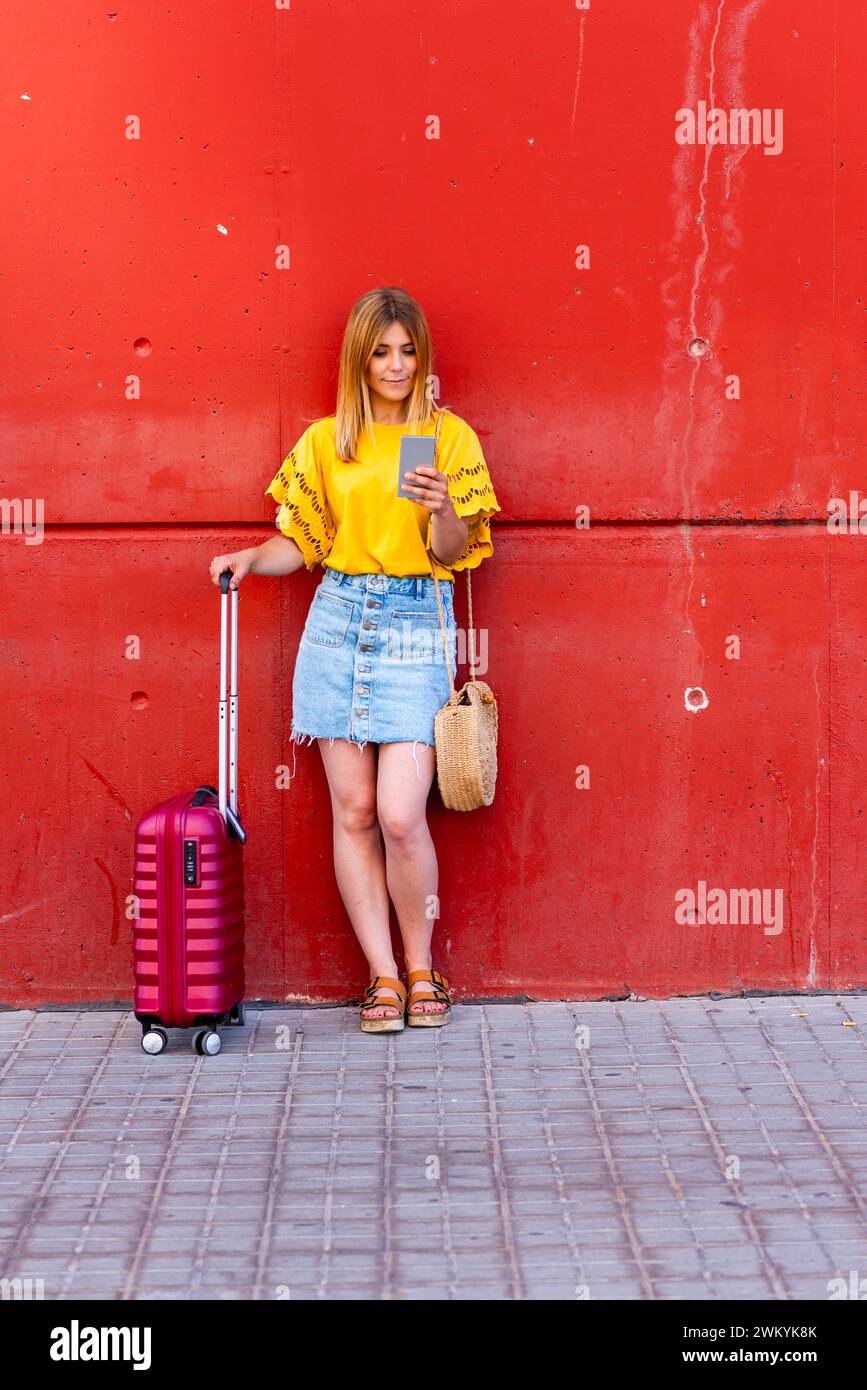 In diesem zeitgenössischen Bild steht eine junge Reisende vor einer markanten roten Wand, ihr rosafarbener Koffer an ihrer Seite. Sie ist mit der digitalen Welt verbunden Stockfoto