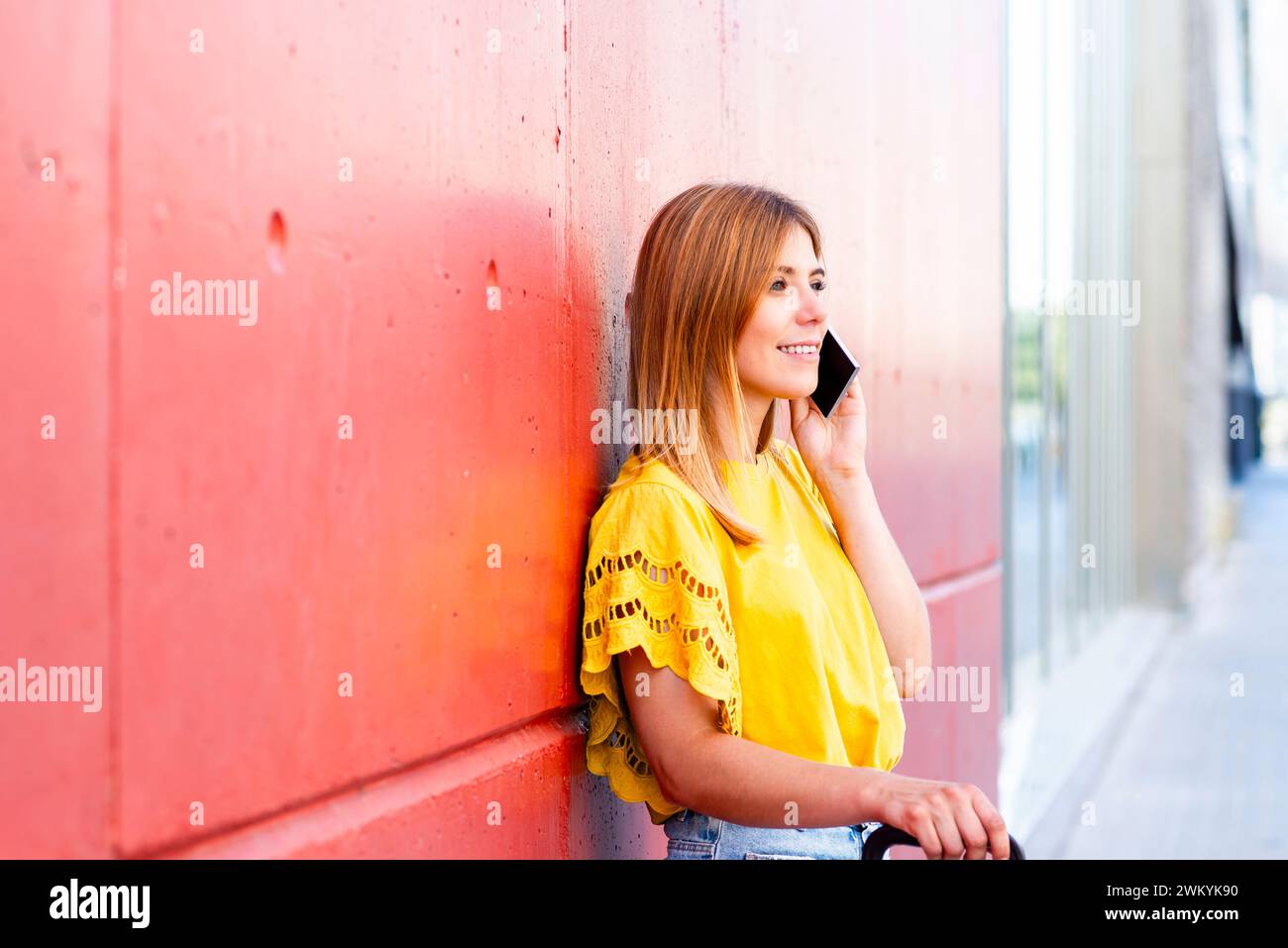 Eine junge Frau lehnt sich lässig an eine strukturierte rote Wand, tief im Gespräch mit ihrem Smartphone. Ihre hellgelbe Bluse und ihr entspanntes Auftreten geben es Stockfoto