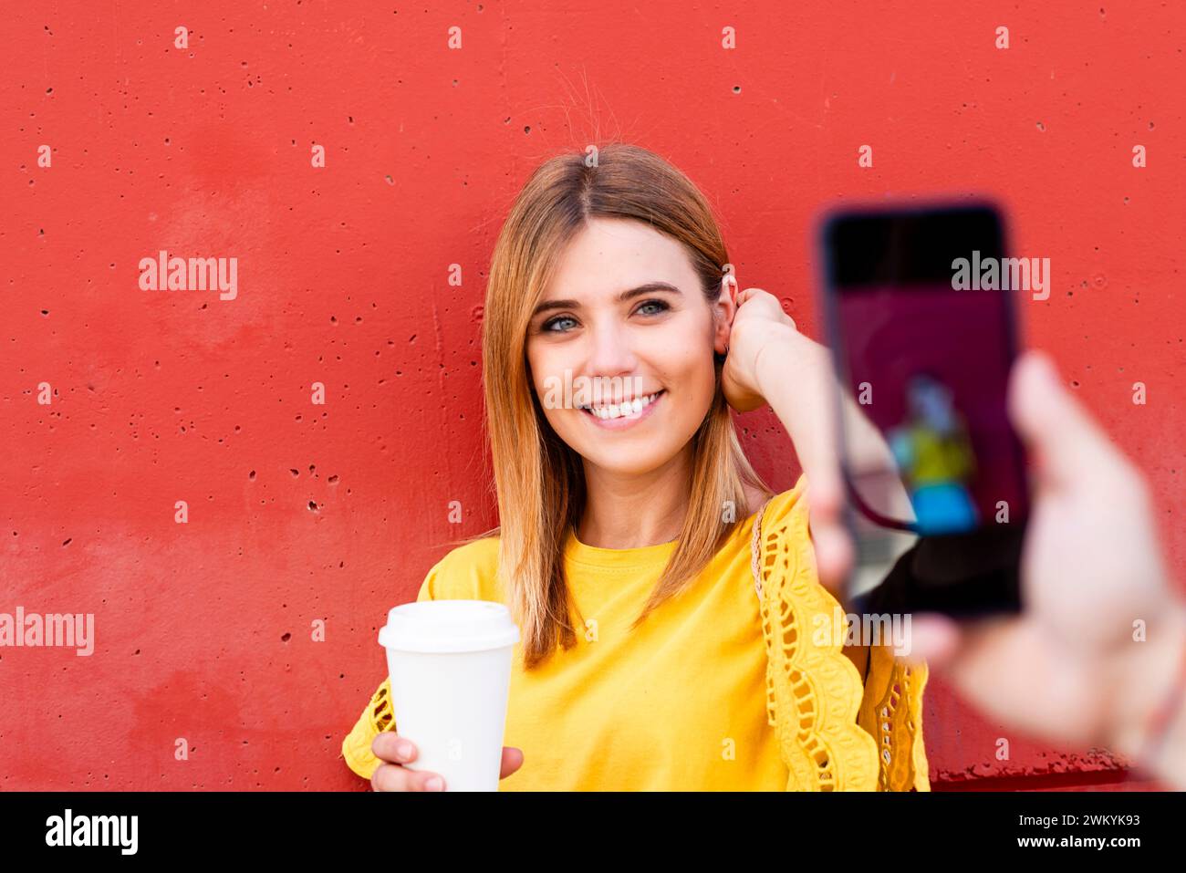 Eine lebhafte Fotosession in der Stadt in Aktion: Eine junge Frau mit einem Kaffee in der Hand blitzt ein helles Lächeln, während sie für ein Smartphone-Foto posiert. Ihr Gelb Stockfoto