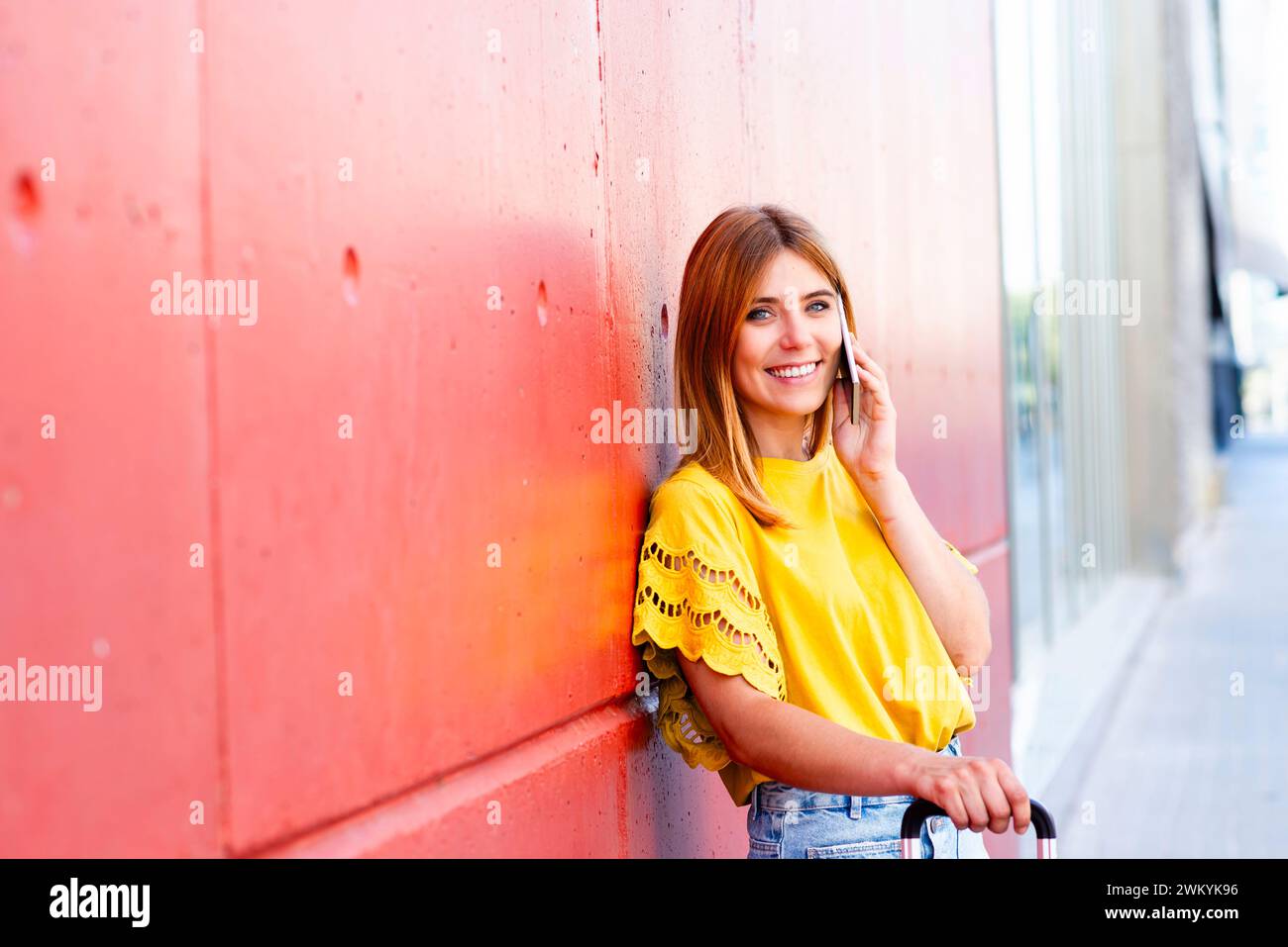 Eine junge Frau lehnt sich lässig an eine strukturierte rote Wand, tief im Gespräch mit ihrem Smartphone. Ihre hellgelbe Bluse und ihr entspanntes Auftreten geben es Stockfoto