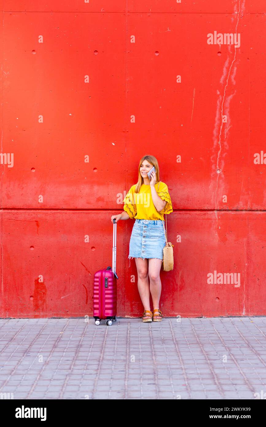 Dieses Foto erzählt die Geschichte eines Moments auf der Durchreise, als eine junge Frau, die vor einer leuchtend roten Wand steht, mit einem rosa Koffer von h auf ihrem Handy spricht Stockfoto
