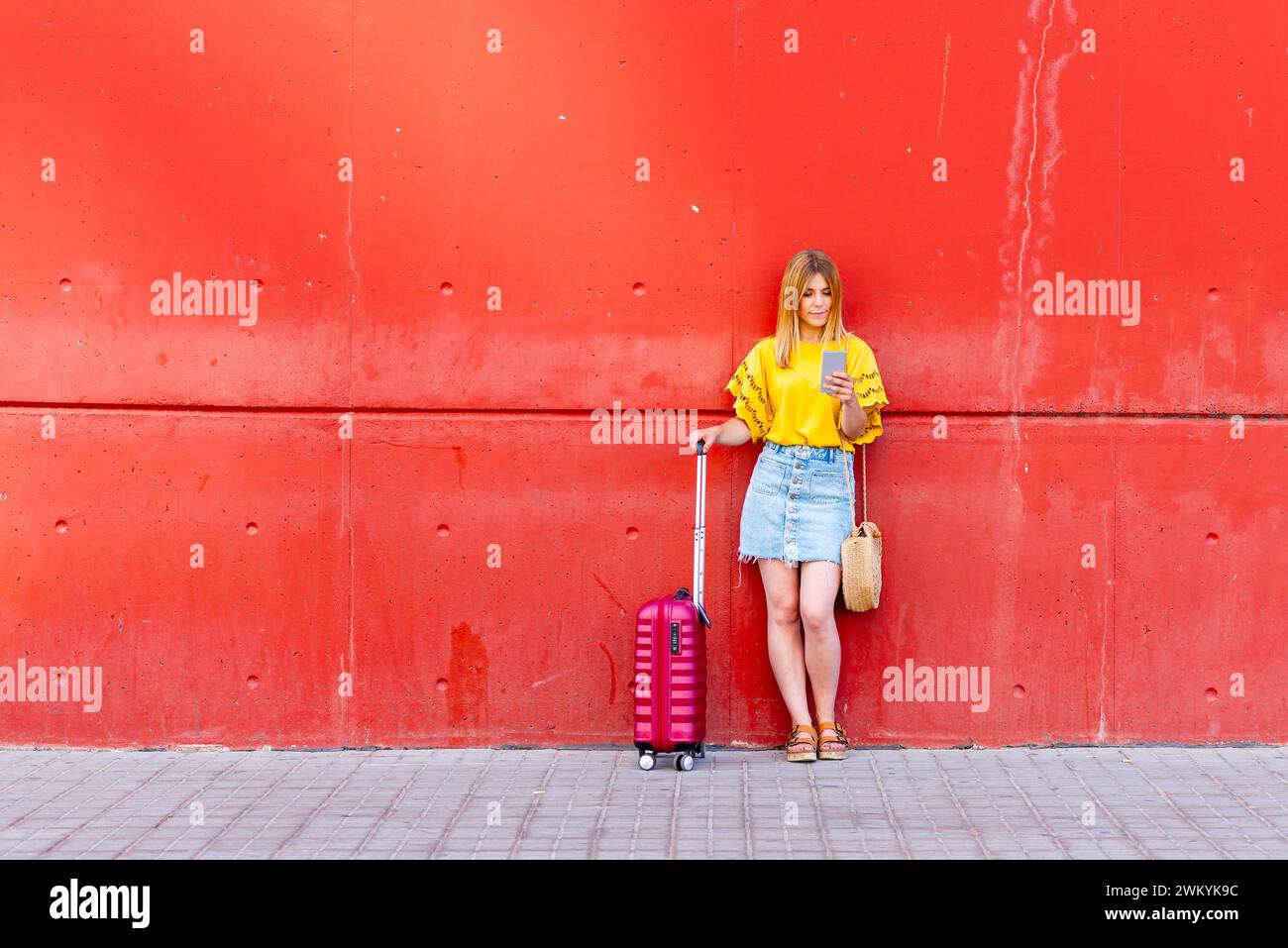 In diesem zeitgenössischen Bild steht eine junge Reisende vor einer markanten roten Wand, ihr rosafarbener Koffer an ihrer Seite. Sie ist mit der digitalen Welt verbunden Stockfoto