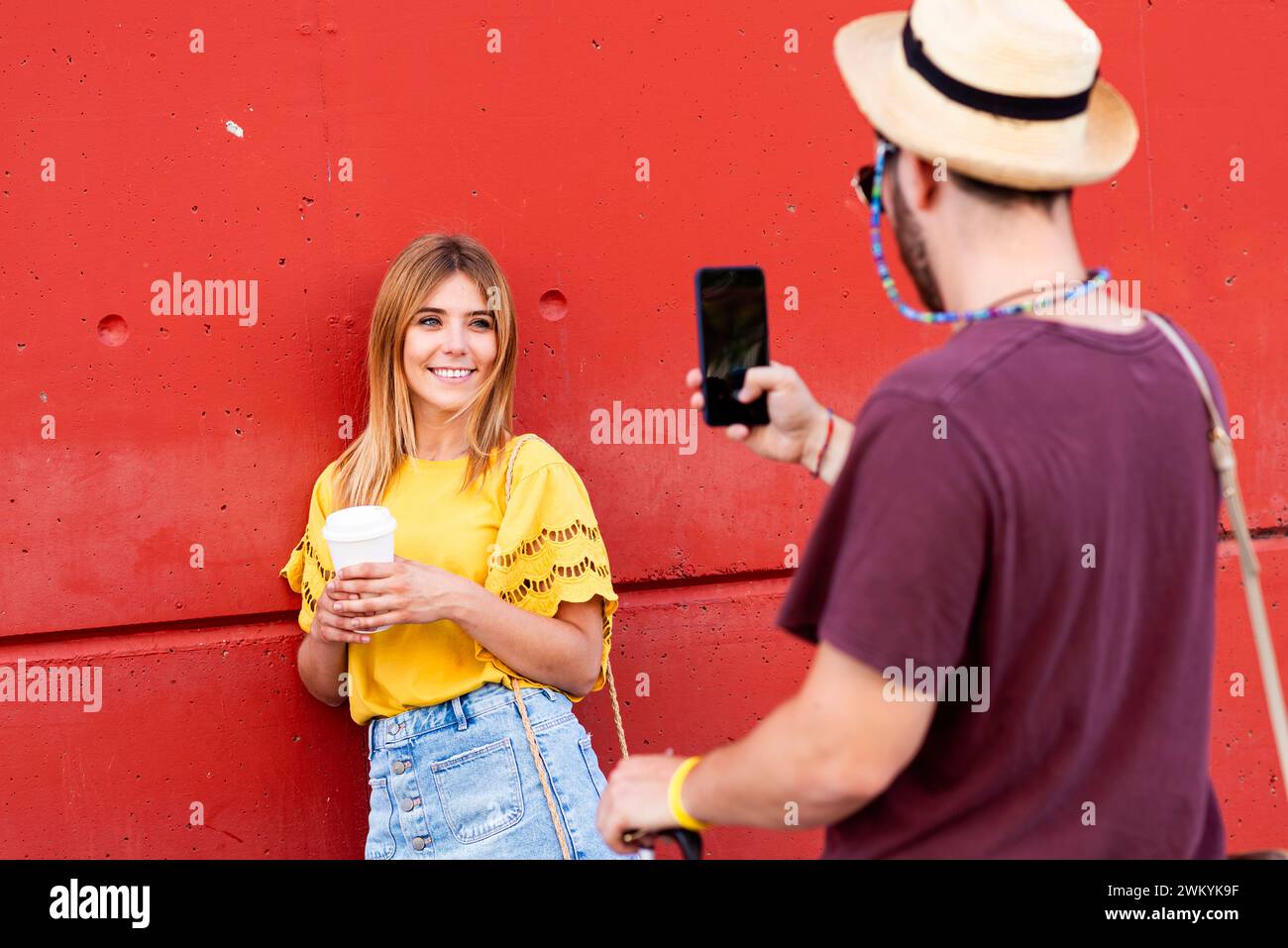 Eine lebhafte Fotosession in der Stadt in Aktion: Eine junge Frau mit einem Kaffee in der Hand blitzt ein helles Lächeln, während sie für ein Smartphone-Foto posiert. Ihr Gelb Stockfoto