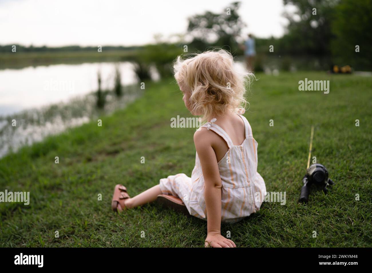 Kleines Mädchen, Das Neben Dem Teich Sitzt Und Einen Angelmast Hat Stockfoto