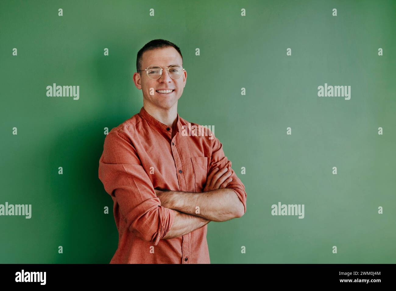 Lächelnder Mann mit Brille, Arme vor grüner Wand gekreuzt Stockfoto