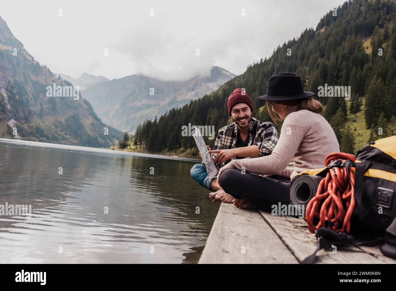 Glückliches junges Paar, das mit Karte auf dem Pier in der Nähe des Vilsalpsees und der Berge in Tirol, Österreich, sitzt Stockfoto