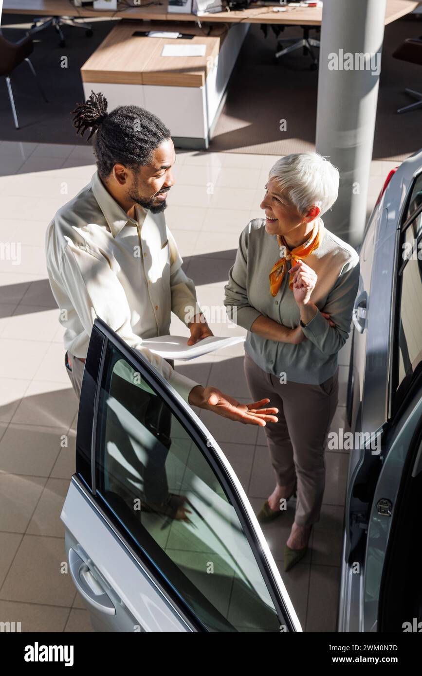 Glückliche Seniorin mit Verkäuferin, die das Auto im Ausstellungsraum zeigt Stockfoto