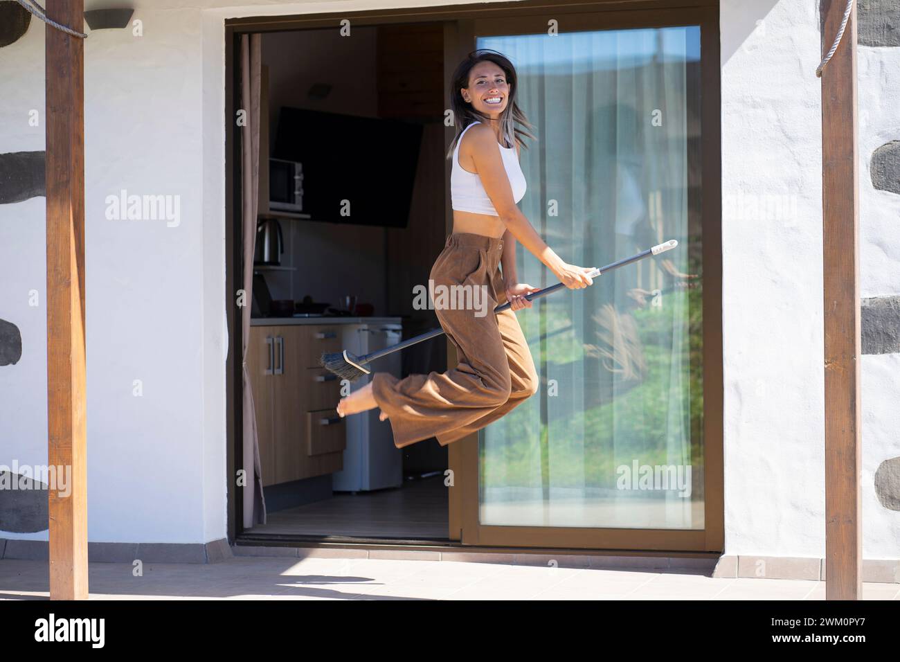 Verspielte Frau mit Besen in der Luft auf der Veranda Stockfoto