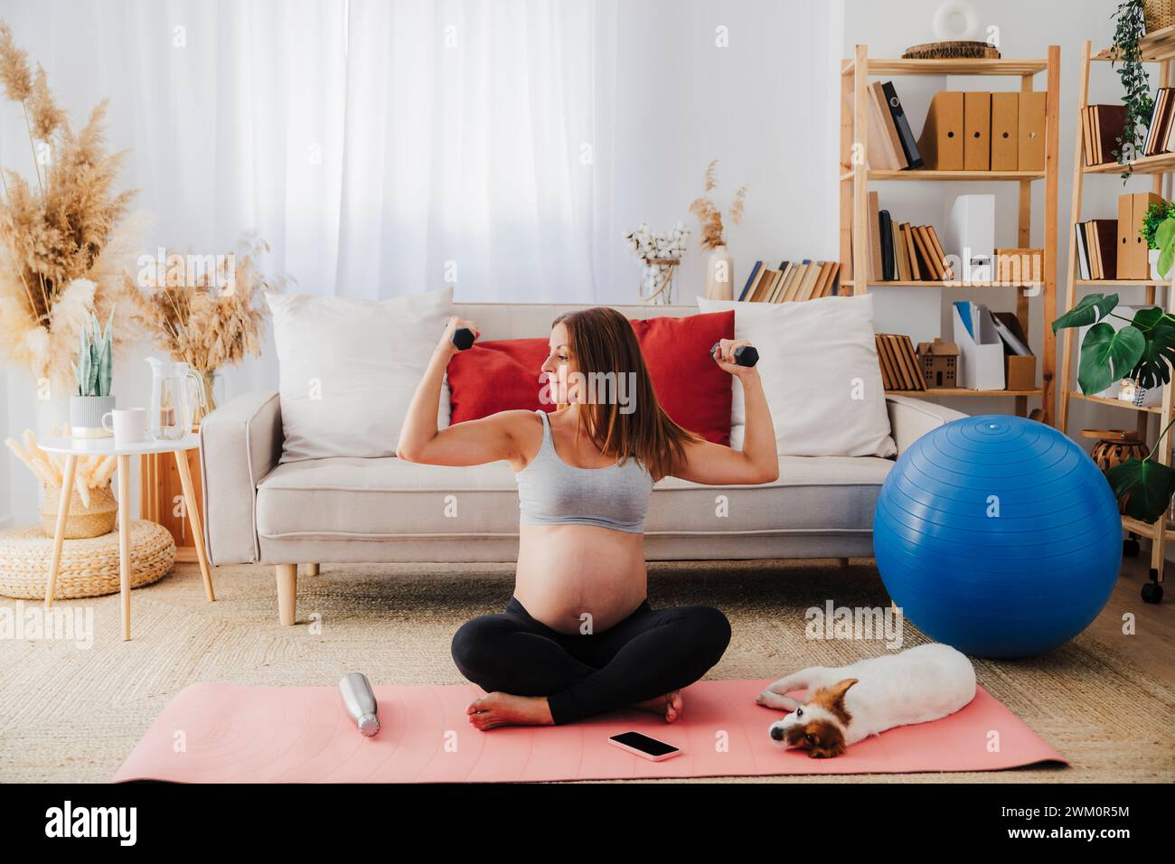 Schwangere Frau, die mit Hanteln zu Hause im Kreuz auf Yogamatte sitzt Stockfoto
