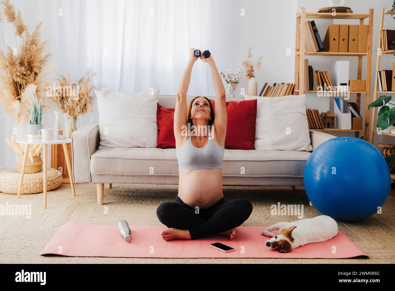 Schwangere Frau, die zu Hause mit Hanteln auf Yogamatte sitzt Stockfoto