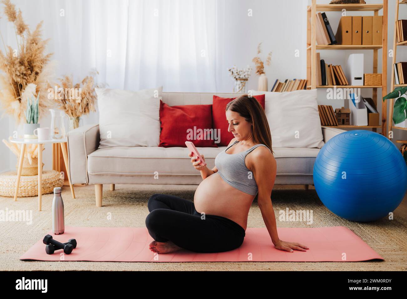Schwangere Frau mit Smartphone auf der Trainingsmatte im Wohnzimmer zu Hause Stockfoto