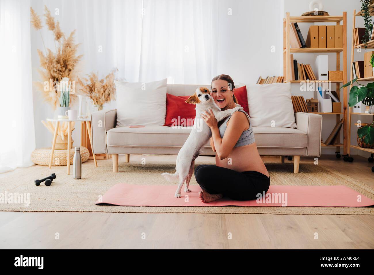 Glückliche schwangere Frau, die mit Hund spielt, der zu Hause auf Yogamatte sitzt Stockfoto