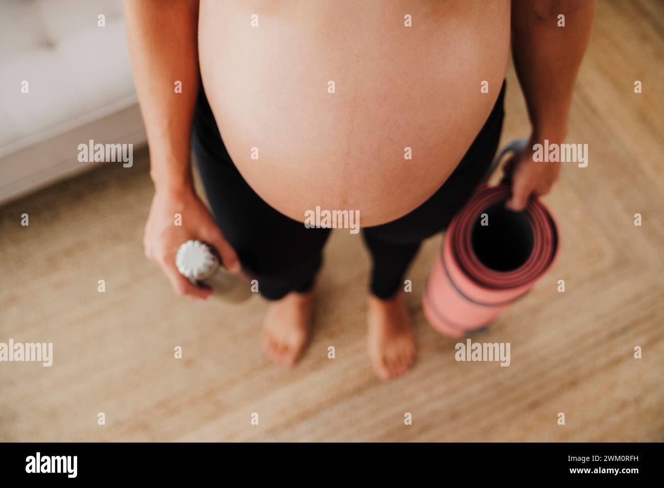 Schwangere Frau, die zu Hause mit Wasserflasche und Yogamatte steht Stockfoto