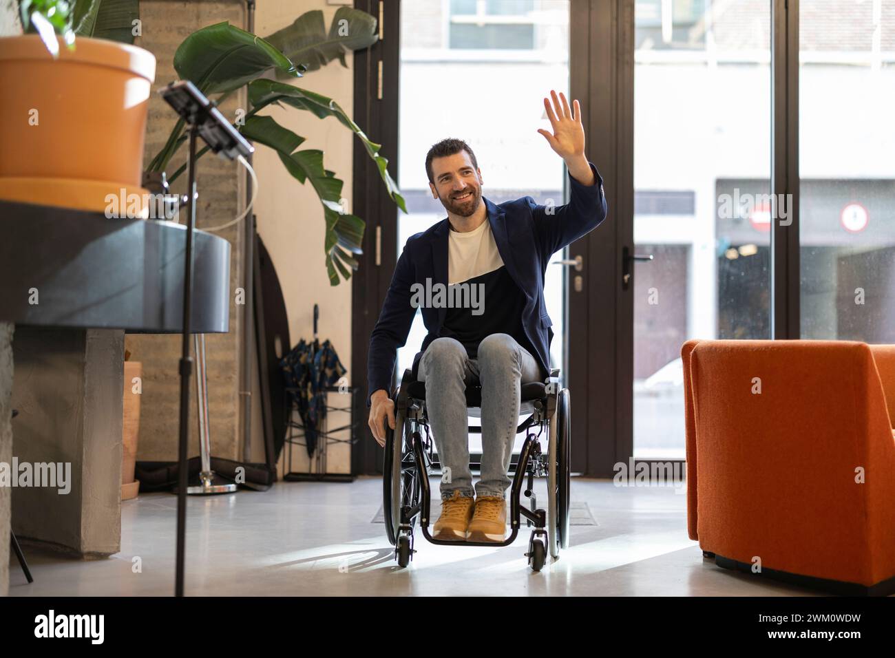 Lächelnder Geschäftsmann im Rollstuhl mit Behinderung, der am Arbeitsplatz winkt Stockfoto