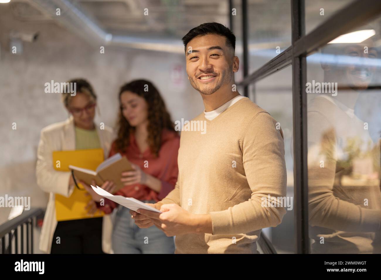 Lächelnder junger Geschäftsmann, der mit einem Dokument neben Kollegen steht, die in einem beleuchteten Korridor diskutieren Stockfoto