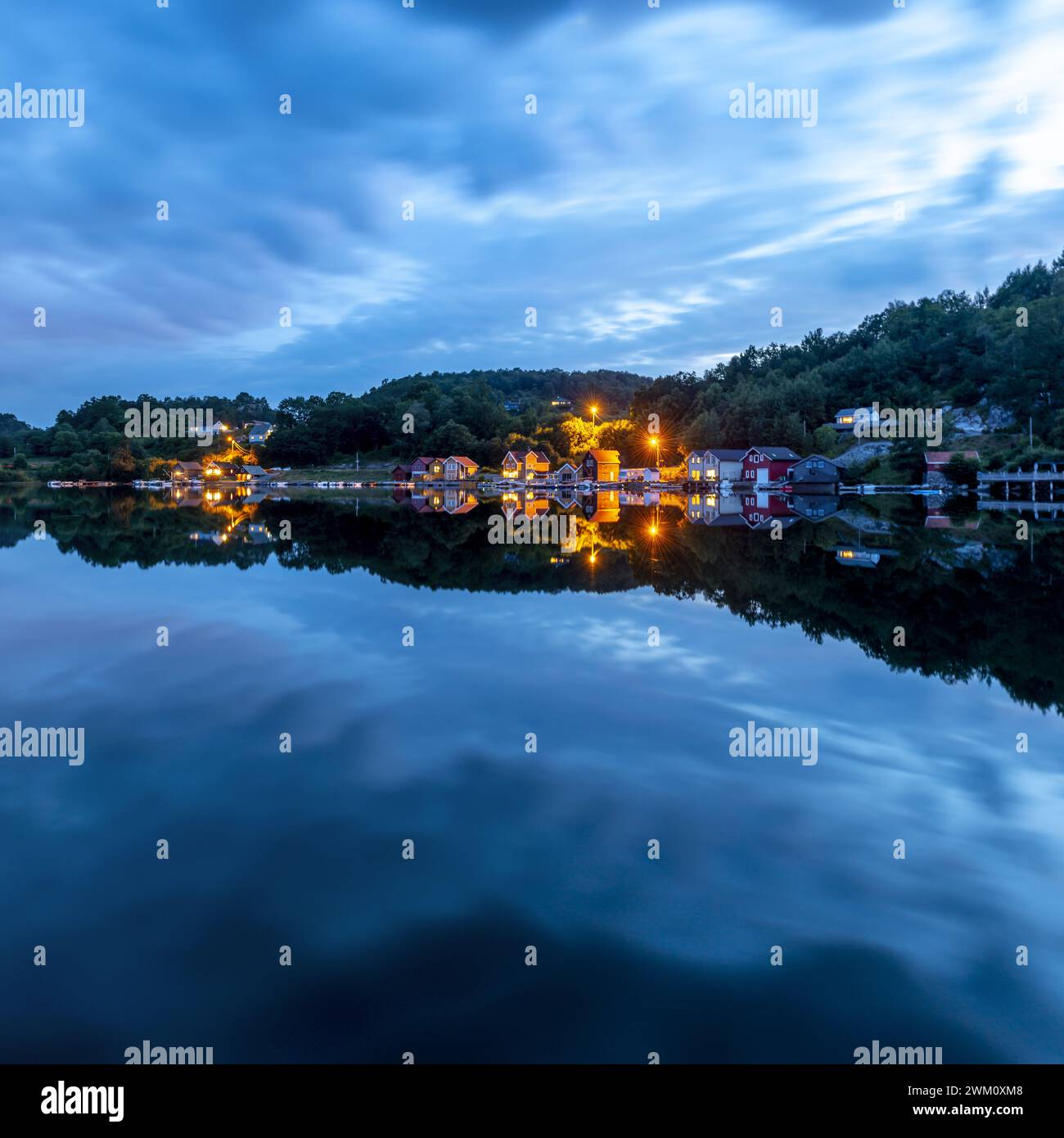 Am Abend spiegeln sich die Lichter der Uferentwicklung in einem spiegelglatten See unter einem blauen, spektakulär bewölkten Himmel. Stockfoto
