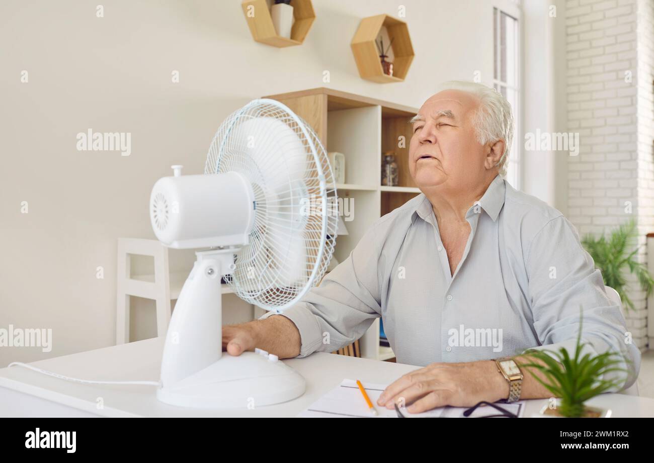 Senior Mann, müde von Sommerhitze, versucht, sich von einem elektrischen Ventilator zu Hause abzukühlen Stockfoto