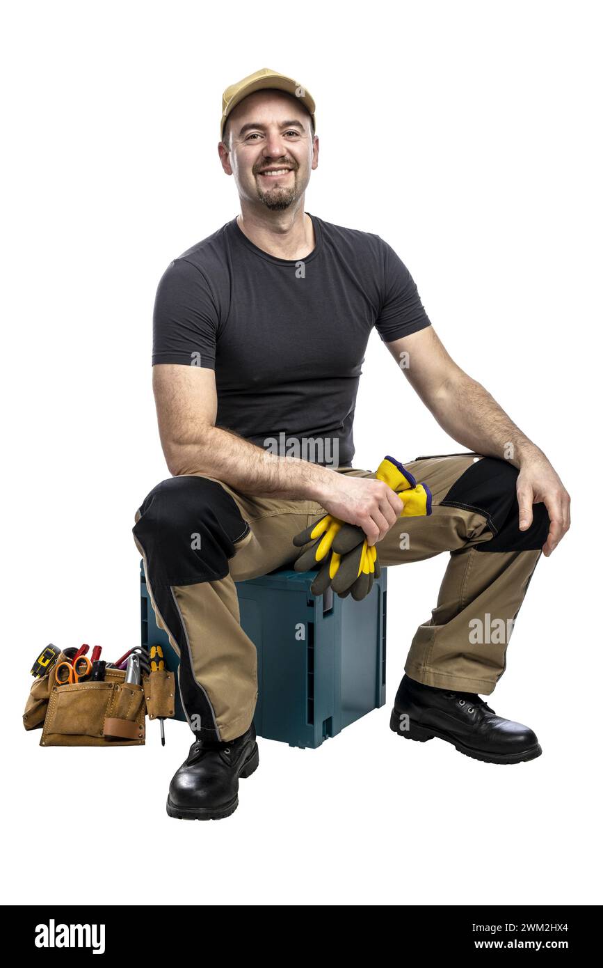 Fröhlicher männlicher Handwerker sitzt mit Werkzeugkasten und Handschuhen, bereit für die Arbeit Stockfoto