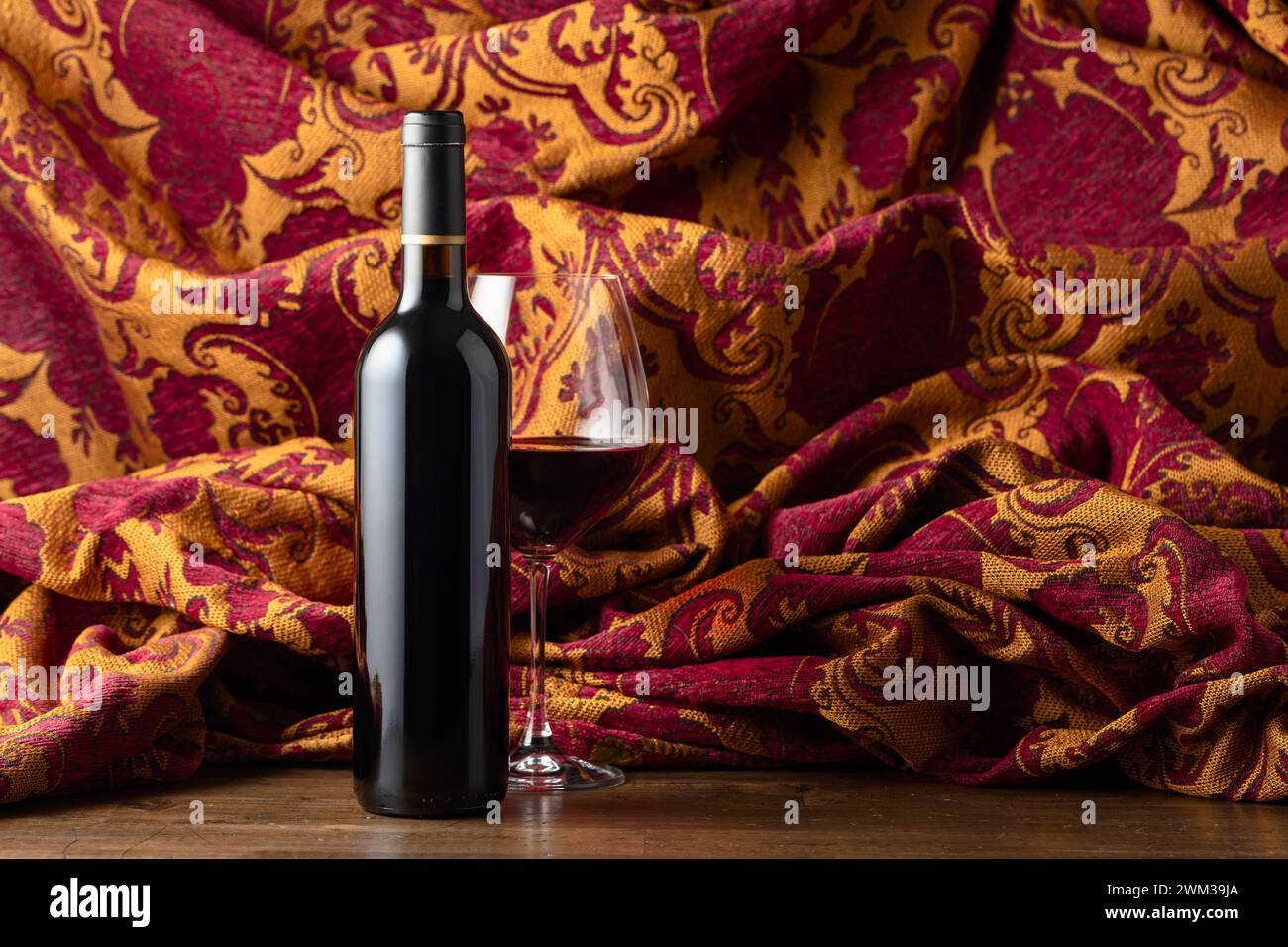 Flasche und Glas Rotwein auf einem alten Holzteller. Im Hintergrund Retro-Wandteppich mit dunkelrotem und goldenem Blumenschmuck. Stockfoto
