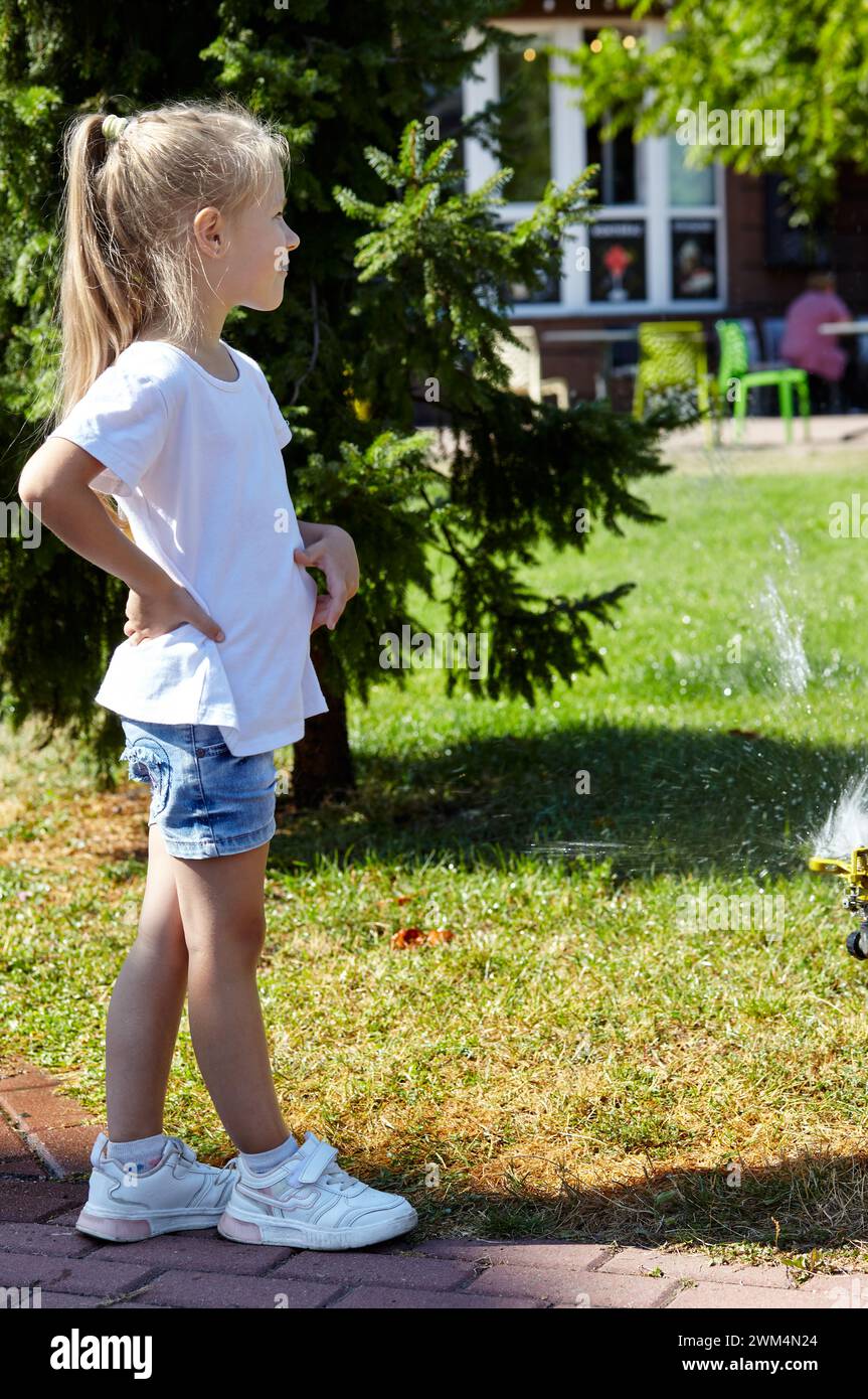 Kleines Mädchen, das unter dem Wasser von der Sprinklerbewässerung im Sommer Stadtpark läuft. Kindheit, Freizeit und Menschenkonzept - glückliche Kinderruhe und Stockfoto