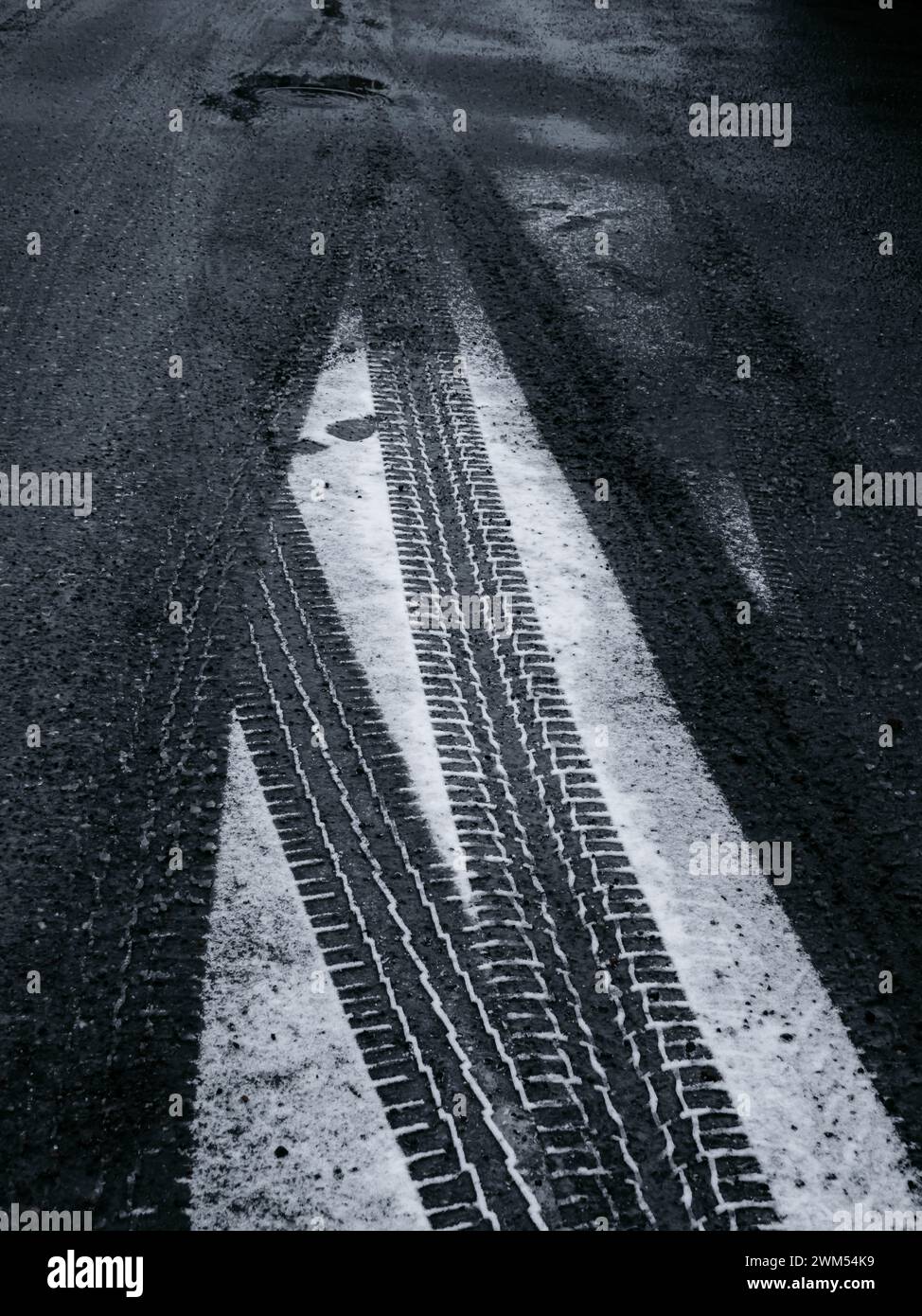 Der dunkle Asphalt einer Straße in Molndal, Schweden, ist teilweise mit Schnee bedeckt und zeigt die kontrastierenden schwarzen Reifenspuren. Am Abend wird das Licht schwächer Stockfoto
