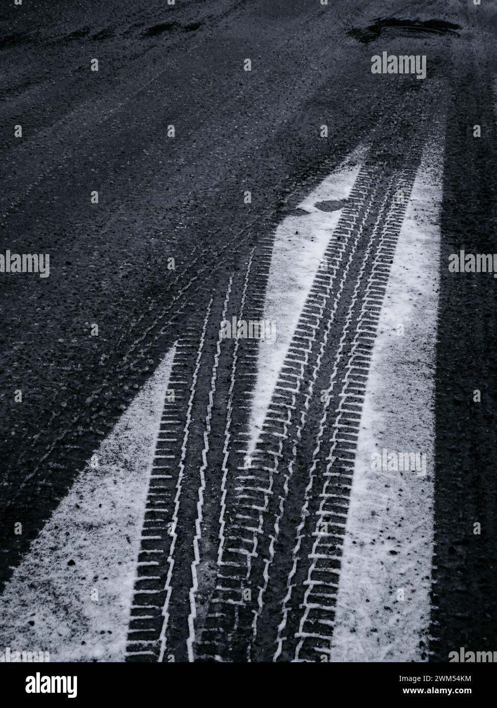 Der dunkle Asphalt einer Straße in Molndal, Schweden, ist teilweise mit Schnee bedeckt und zeigt die kontrastierenden schwarzen Reifenspuren. Am Abend wird das Licht schwächer Stockfoto