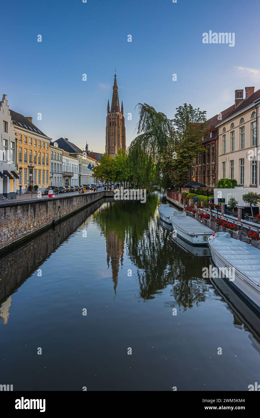 Abendliche Atmosphäre auf einem Kanal in der Altstadt von Brügge. Alte Gebäude und Boote entlang des Wassers mit einer Straße. Im Hintergrund steht die Kirche Stockfoto