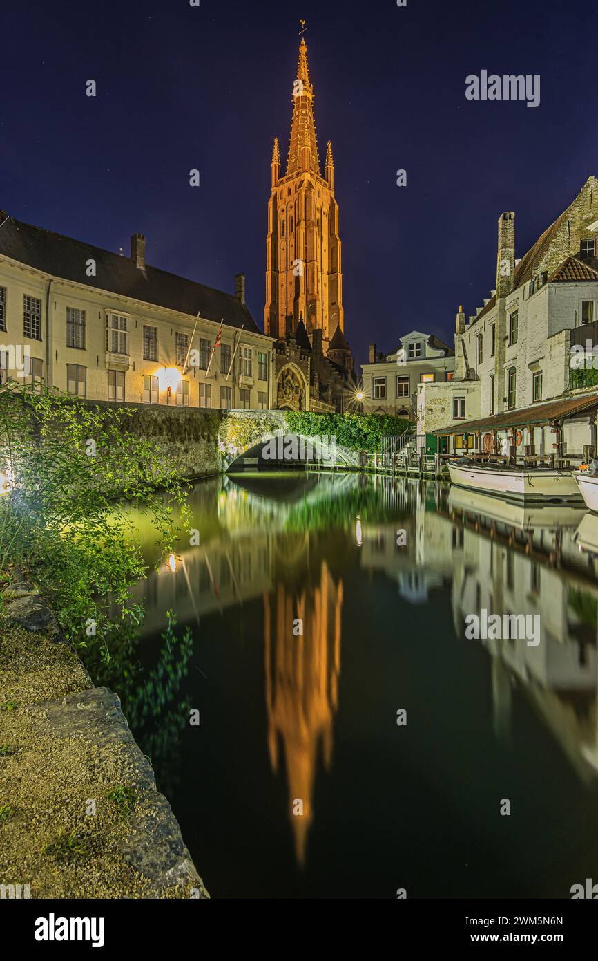 Kanal in der belgischen Hansestadt Brügge am Abend. Kirche unserer Lieben Frau und beleuchtete historische Gebäude zur blauen Stunde. Reflexionen auf Wasser Stockfoto
