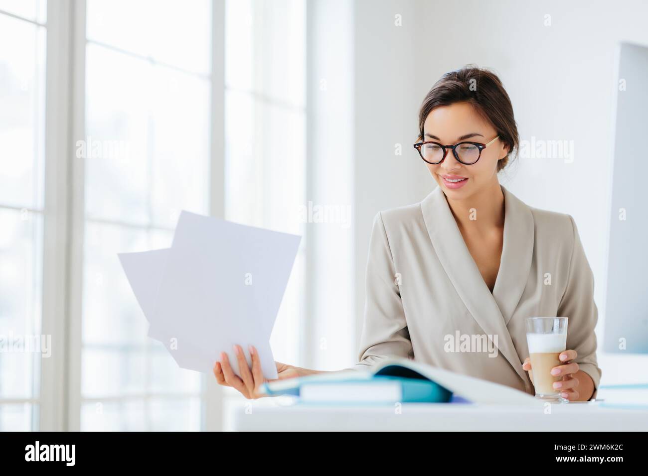 Geschäftsfrau im schicken beigefarbenen Blazer hält Papiere mit einem Glas Wasser, gesundheitsbewusst. Stockfoto