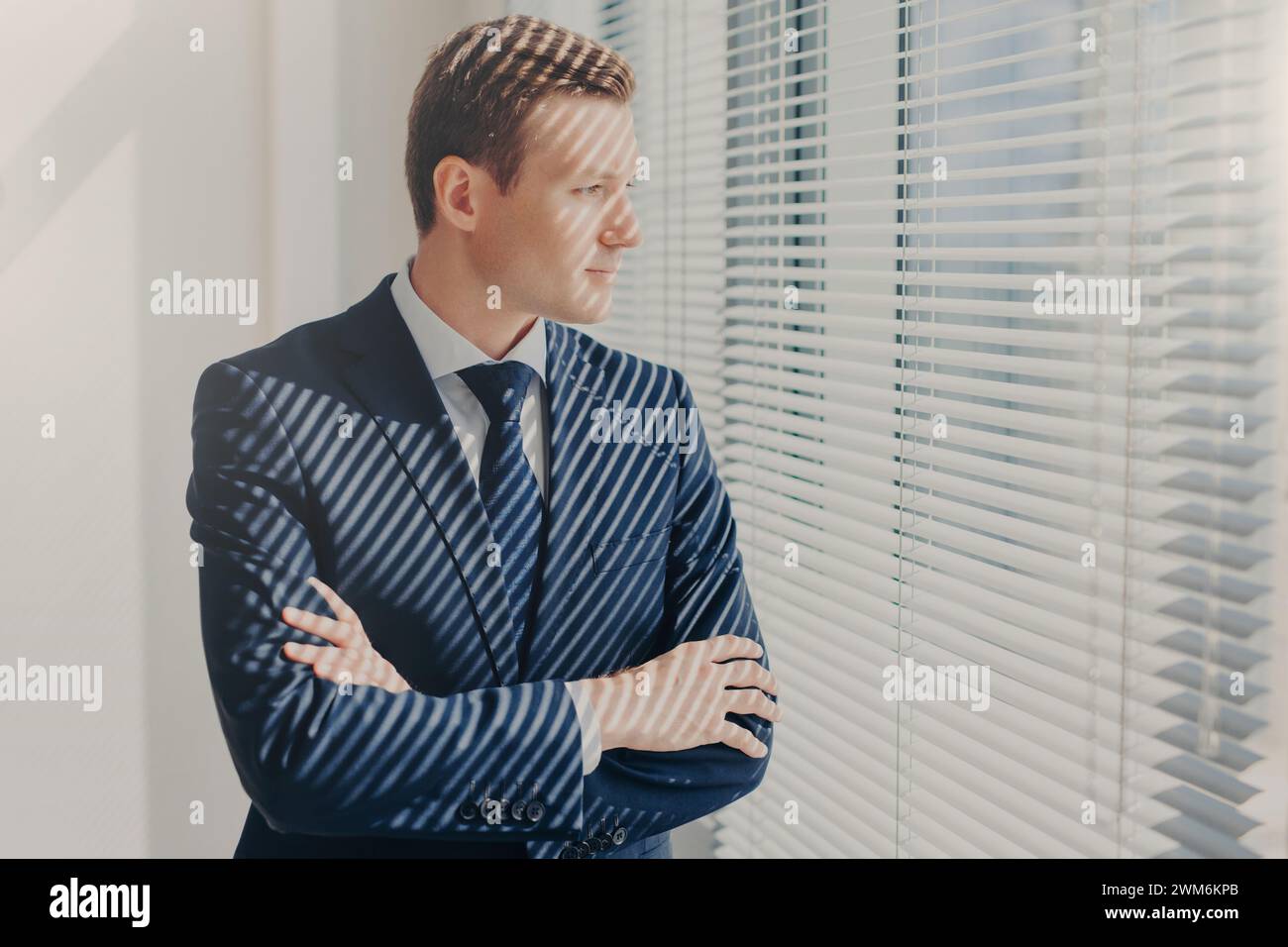 Reflektierender Geschäftsmann mit überkreuzten Armen, der mit einem nachdenklichen Ausdruck durch Bürojalousien blickt. Stockfoto