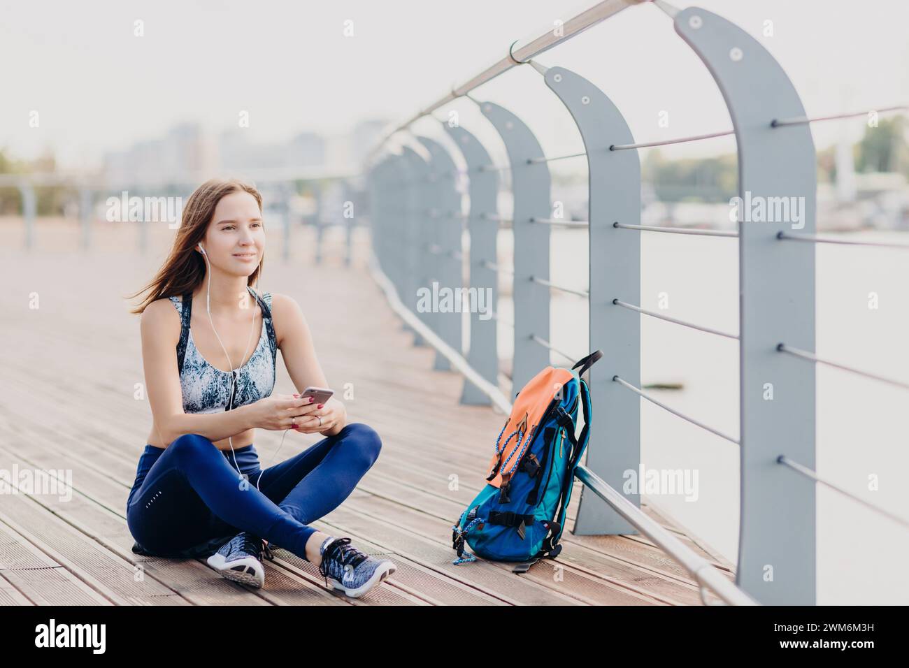 Eine junge Frau mit blauer Aktivkleidung, die am Ufer sitzt und mit ihrem Handy verlobt ist. Stockfoto