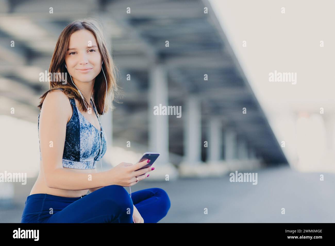Eine lächelnde junge Frau, die unter einer Brücke sitzt und ein Smartphone hält. Stockfoto