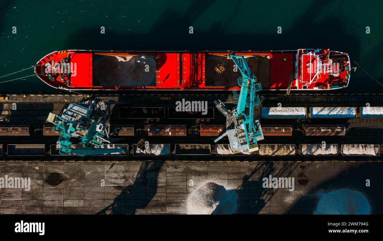 Top-down-Ansicht eines Massengutschiffs, der mit Rohstoffen an einem Industriestandort beladen wird. Das Deck des Schiffes ist offen und zeigt den riesigen Frachtraum, während schwere Maschinen den Transfer erleichtern. Stockfoto