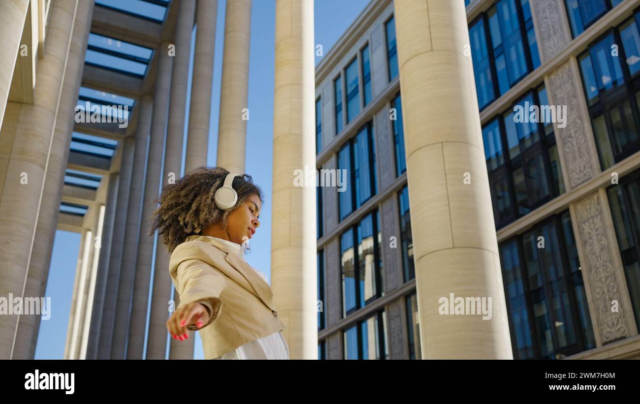 Mit Kopfhörern und einem strahlenden Lächeln tanzt eine junge Berufsfrau frei in einem urbanen Raum, ihre dynamische Bewegung kontrastiert mit den statischen Linien der umgebenden Architektur. Stockfoto