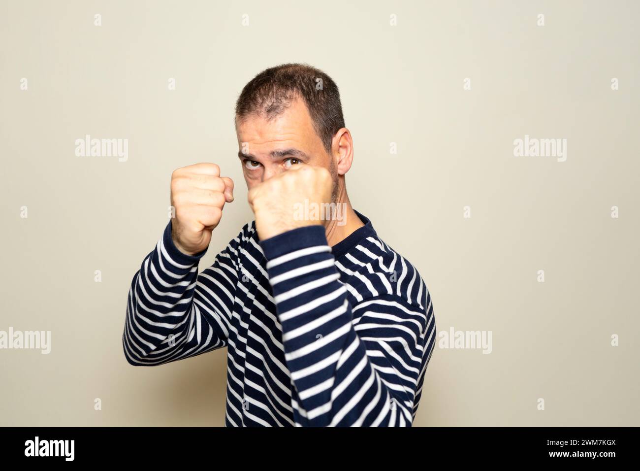 Bärtiger hispanischer Mann in seinen 40ern, der einen gestreiften Pullover trug, mit Fäusten in Verteidigungsposition, bereit zum Kampf, isoliert auf beigefarbenem isoliertem Hintergrund. Stockfoto