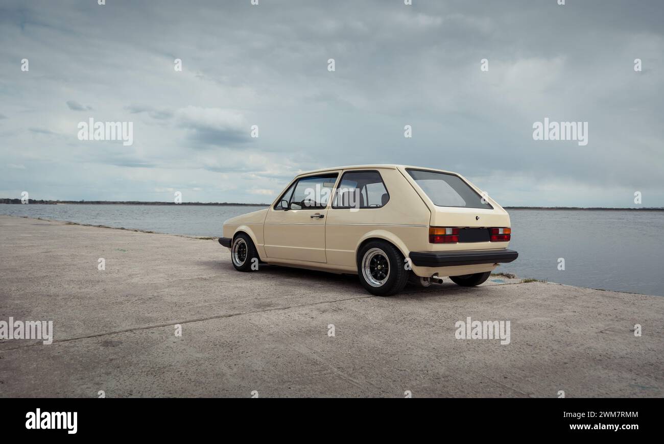 Beigefarbener klassischer Volkswagen Golf auf Betonoberfläche geparkt. Fließheck in der Nähe des Sees an einem bewölkten Tag. Stockfoto