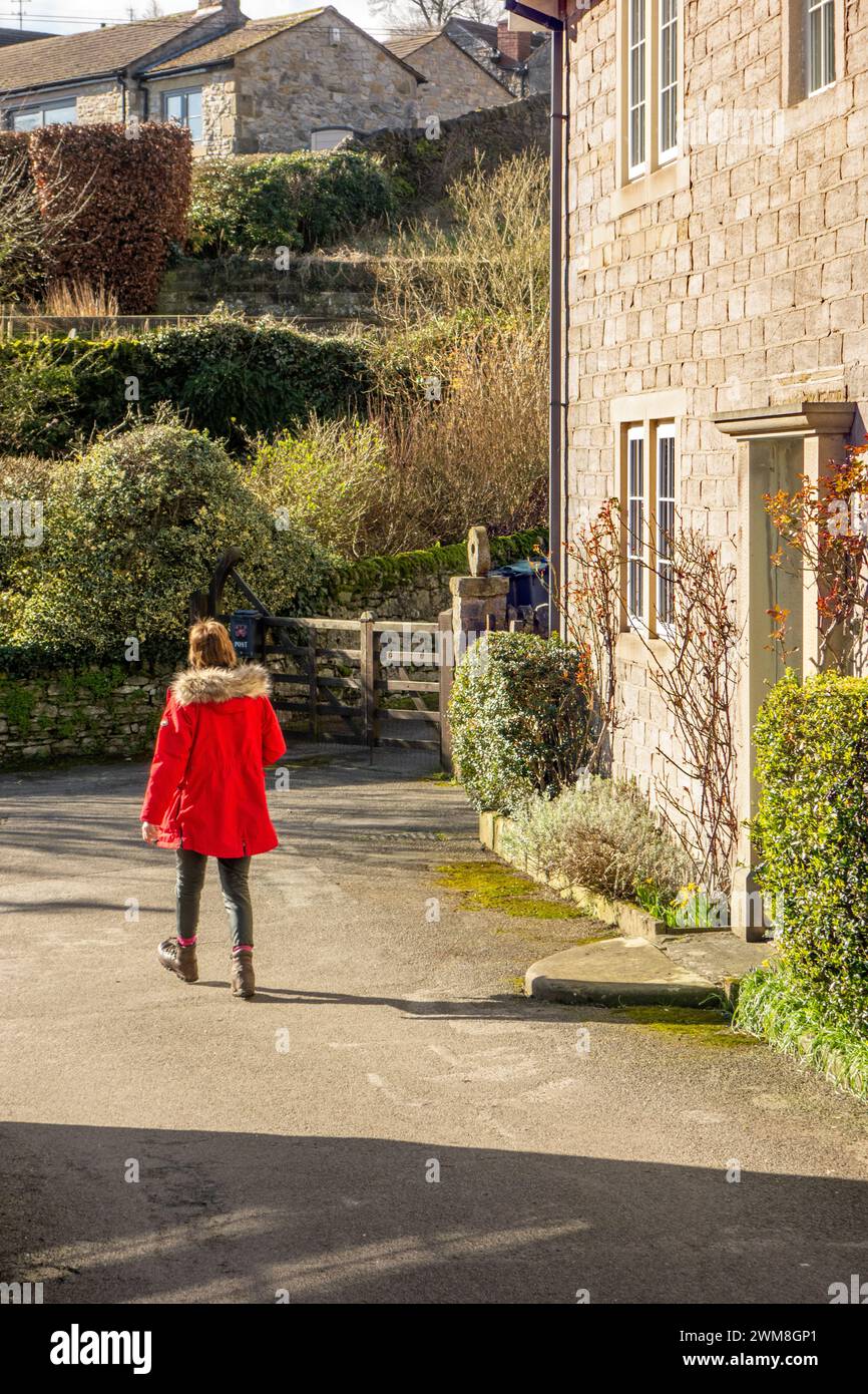 Frau in rotem Mantel, die durch das Peak District Dorf Stoney Middleton läuft, vorbei an Landhäusern Stockfoto
