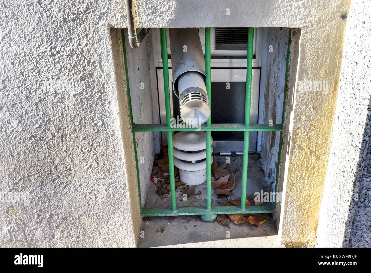 Ein kleines Fenster mit grünem Metallgitter, Aluminiumverschlüssen, trockenen Blättern und Rauchabzugsklappen Stockfoto