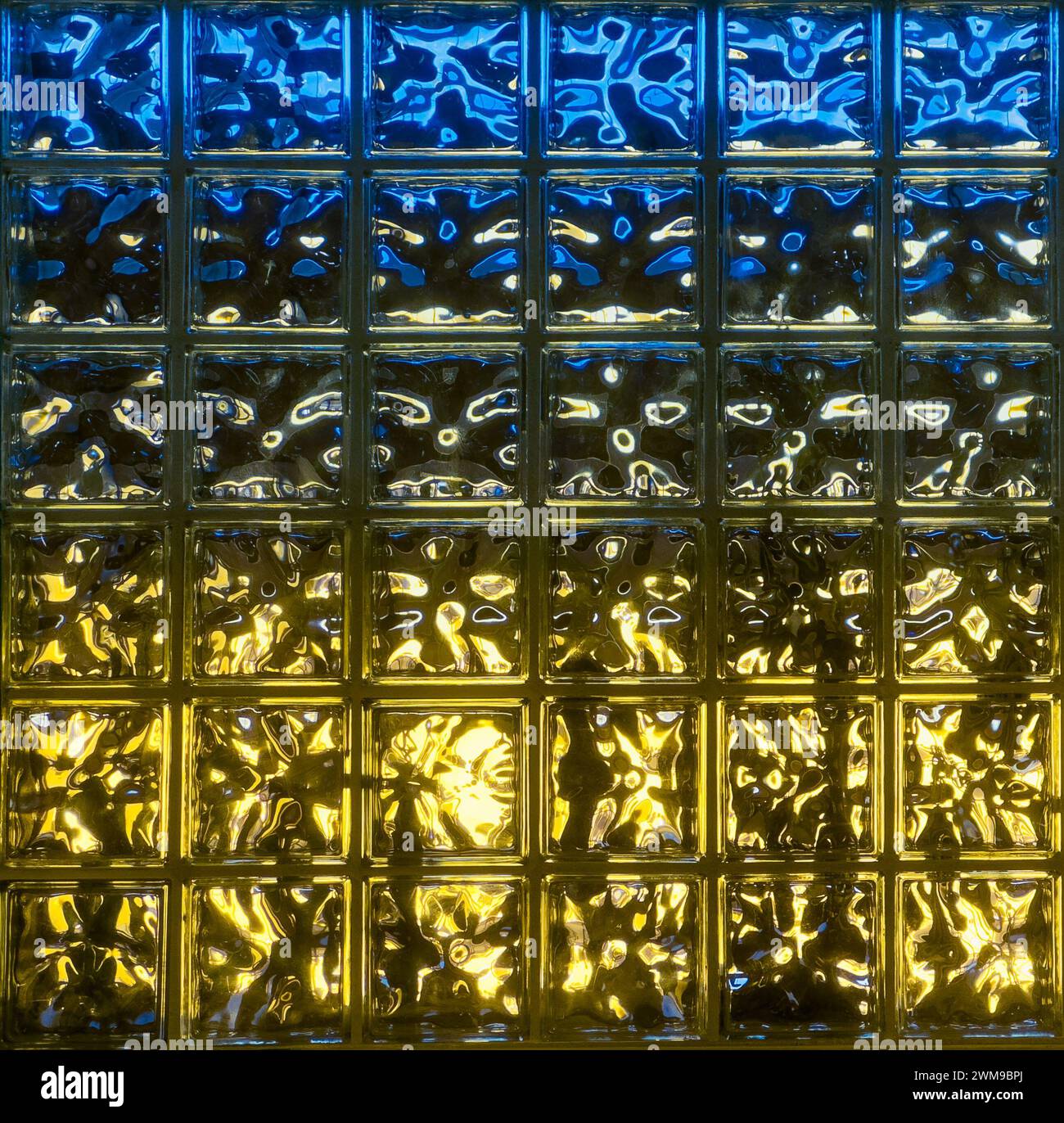 Das Licht, das durch eine Wand aus Glasblöcken hindurchgeht, erzeugt ein farbenfrohes blaues und gelbes wavey-Muster. Stockfoto