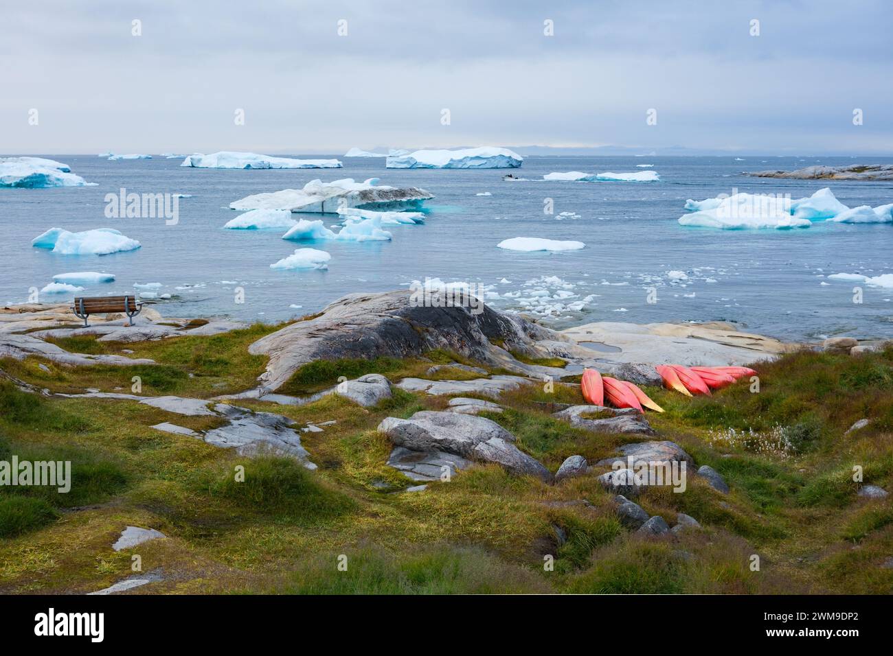 Ein Ort zum Ausruhen: Eine Bank mit Blick auf die Eisberge, die am Ufer schwimmen, und eine Ansammlung roter Kajaks. Illulisat, Grönland. Stockfoto
