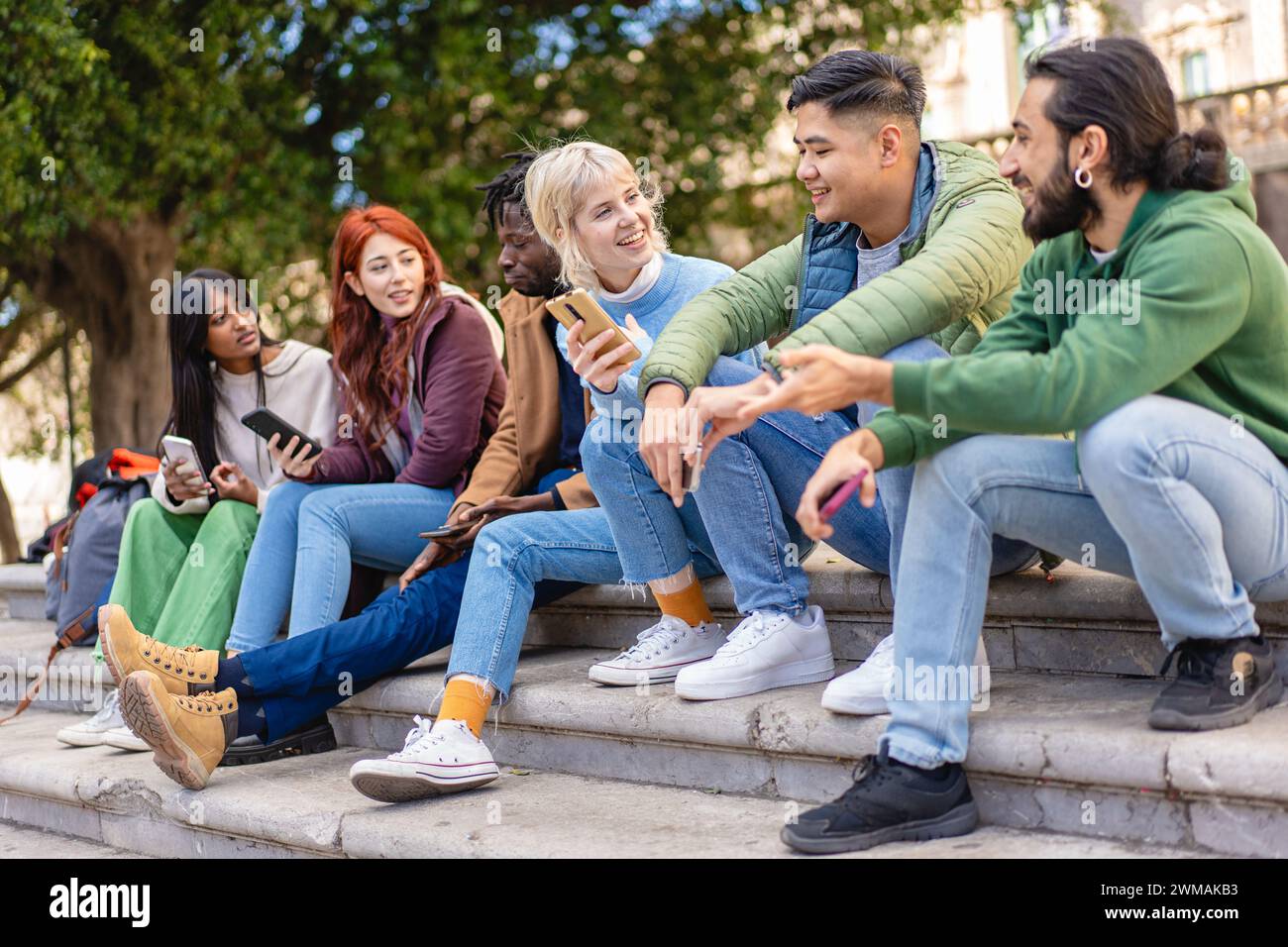 Verschiedene Freunde teilen und lachen über Inhalte auf Smartphones und sitzen auf Stufen in einer urbanen Umgebung. Stockfoto