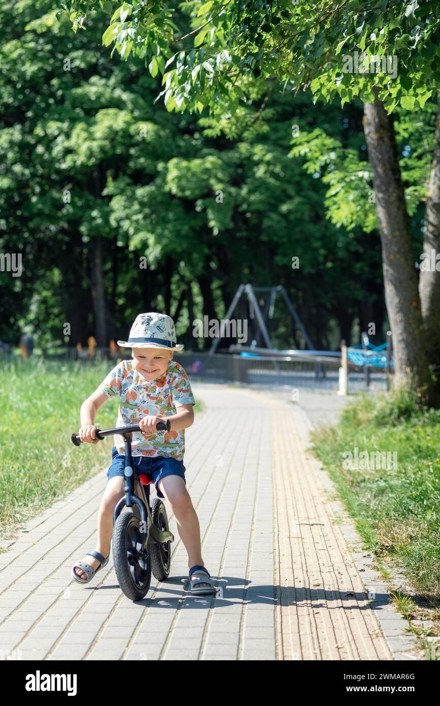 Ein fröhlicher kleiner Junge fährt auf einer Parkanlage in einem Sommerpark auf einem Balance Bike. Stockfoto